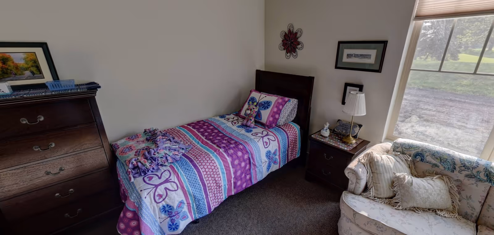 A small bedroom with a single bed covered in a colorful butterfly-themed quilt and a stuffed caterpillar toy on the pillow. Next to the bed is a wooden nightstand with a lamp, decorative items, and framed pictures on the wall. A floral-patterned couch with cushions is positioned near a large window showing a green outdoor scene. A wooden dresser with framed pictures is also visible.