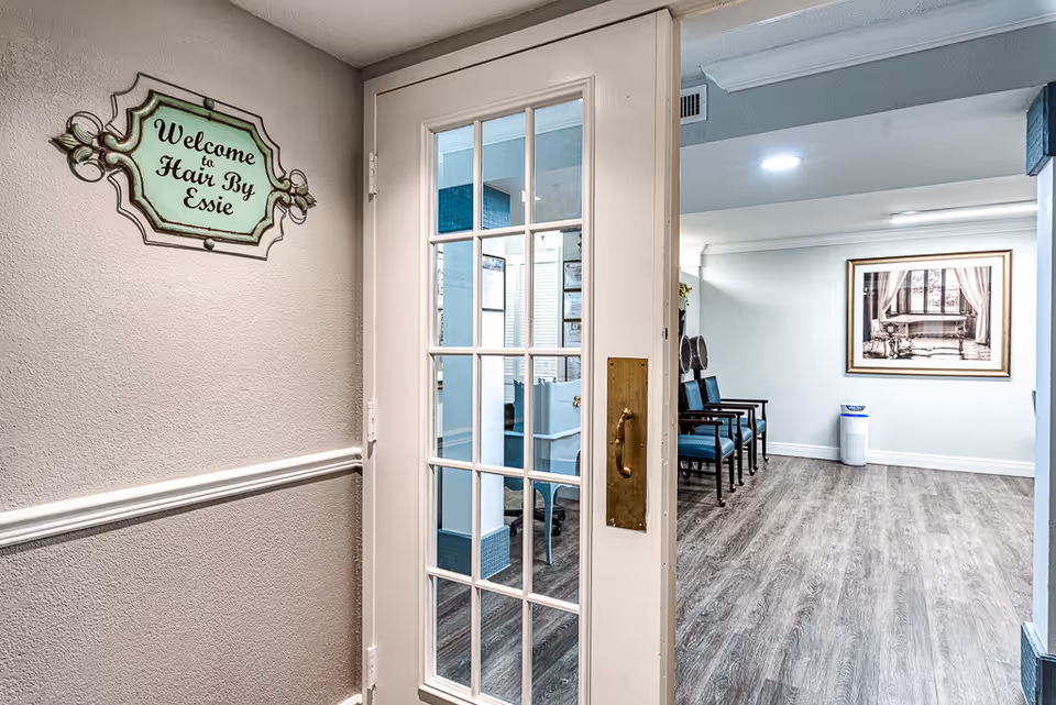 Interior view of a senior living facility hallway leading to a hair salon named 'Hair By Essie'. The hallway has light-colored walls, wood flooring, and a glass-paneled door. Inside the salon, there are chairs lined up against the wall and a framed picture hanging on the far wall.