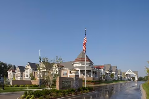 Exterior view of a senior living facility named Waltonwood Main, featuring a long building with multiple peaked roofs, an American flag on a flagpole in front, a paved driveway, and landscaped greenery under a clear blue sky.