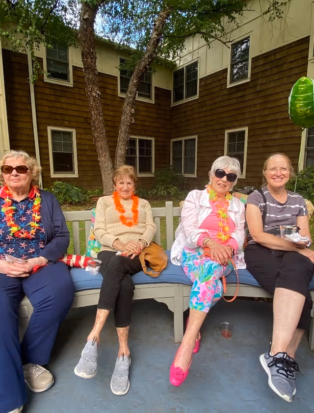Four elderly women sitting on a curved outdoor bench in a courtyard area. They are wearing casual clothing and colorful leis, with one woman wearing sunglasses and bright pink shoes. Behind them is a building with brown siding and several windows, along with some greenery and a tree.