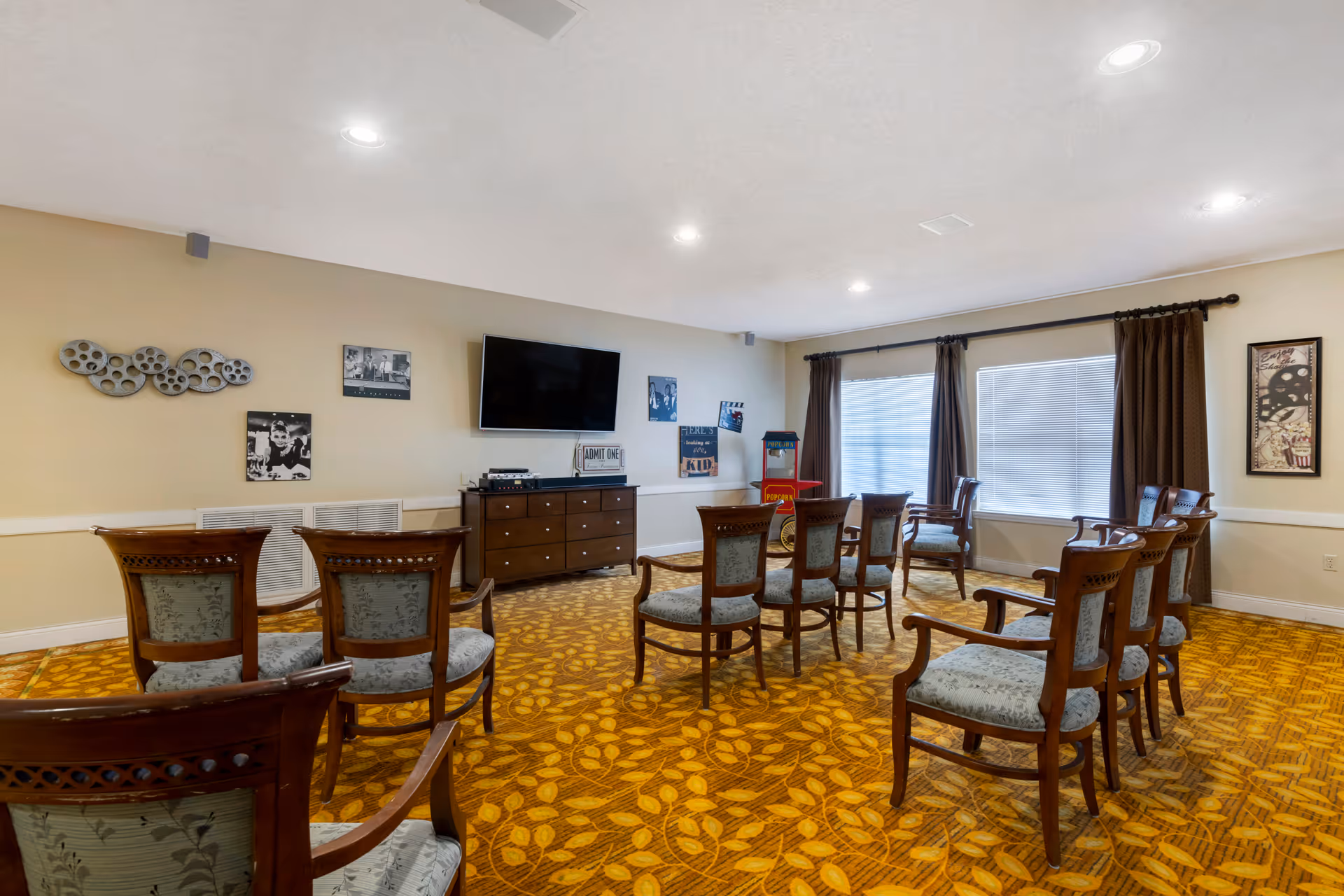 A cozy common room with multiple wooden chairs arranged in rows facing a wall-mounted flat screen TV. The room has a patterned yellow carpet, beige walls decorated with black and white movie-themed pictures, and a popcorn machine near the window with brown curtains.