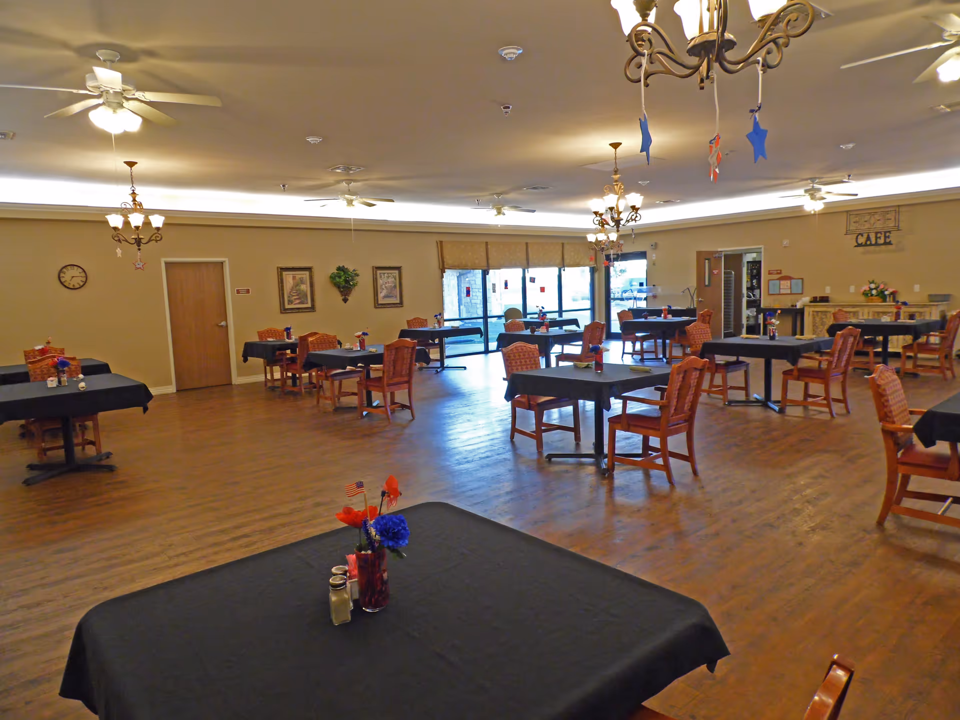 A spacious dining room with multiple tables covered in black tablecloths, each surrounded by wooden chairs with patterned cushions. The room has wooden flooring, ceiling fans, chandeliers, and large windows letting in natural light. There are decorations on the tables and walls, including framed pictures and a clock.