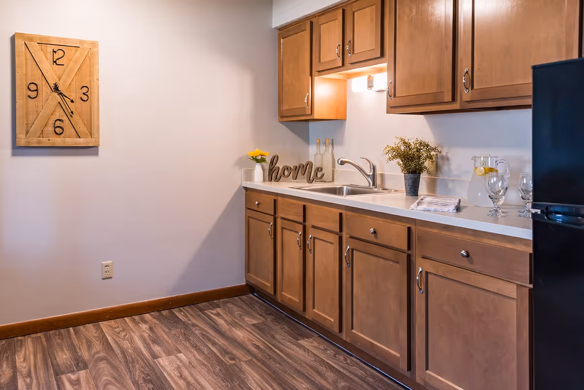 A kitchen area with wooden cabinets, a countertop with a sink, a small potted plant, a decorative 'home' sign, a pitcher of water with lemon slices, and three glasses. A wooden wall clock is mounted on the adjacent wall above a power outlet. The floor has a wood-like finish.