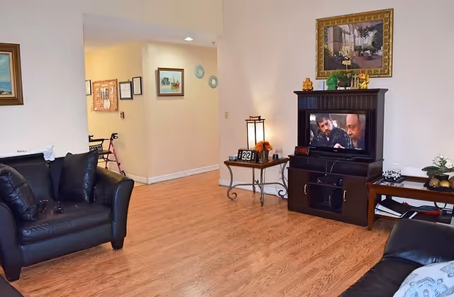 A cozy living room with wooden flooring, two black leather sofas, a dark wooden TV stand with a television showing two men, a side table with a lamp and decorative items, and framed artwork on the walls.