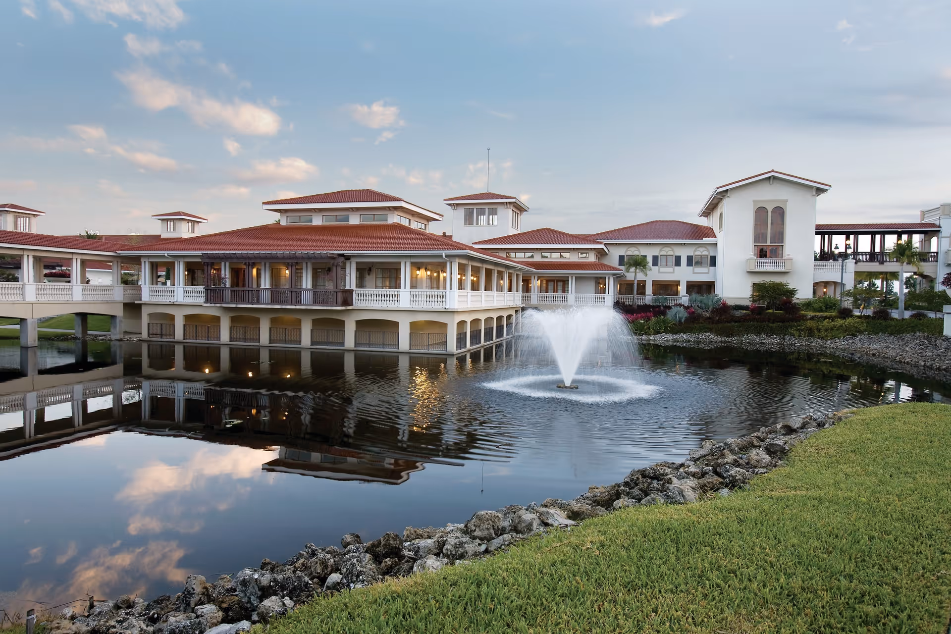 Exterior view of a senior living facility named La Posada with a large pond featuring a water fountain in the center. The building has a red-tiled roof, white walls, and multiple windows. The sky is partly cloudy and the pond reflects the building and sky. There is a grassy area with rocks bordering the pond.