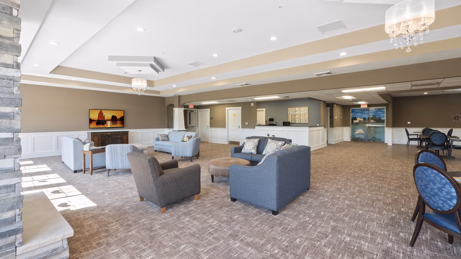 A spacious and well-lit living room area in an assisted living facility featuring multiple seating arrangements with sofas and armchairs, a stone fireplace on the left, a flat-screen TV mounted on the far wall, and a reception desk in the background. The room has beige walls with white wainscoting, carpeted floors, and ceiling lights with chandeliers.