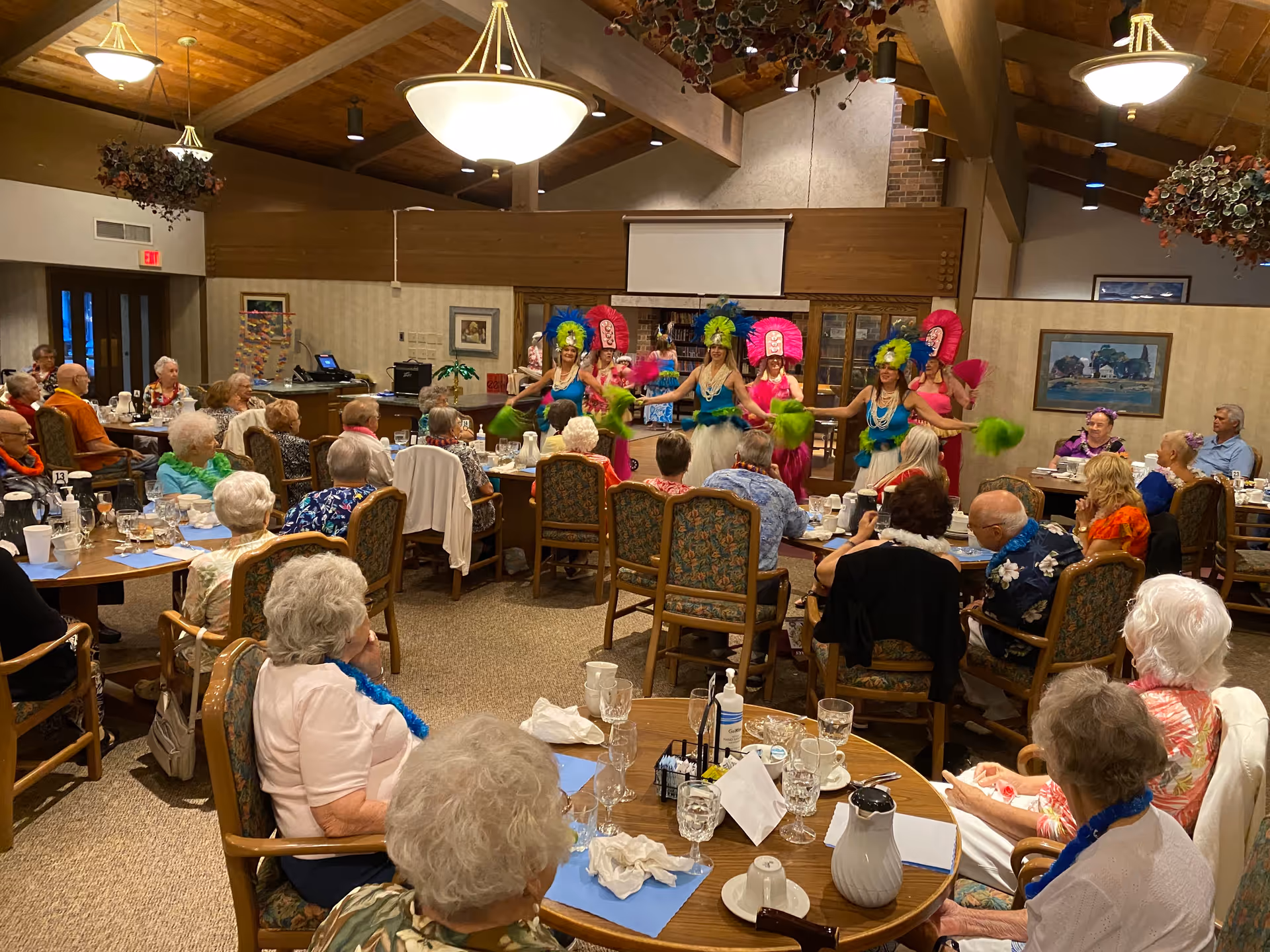 A group of elderly people seated at round dining tables watching a lively performance by dancers dressed in colorful Hawaiian-style costumes with feathered headdresses and fans in a community dining room with wooden ceiling beams and hanging plants.