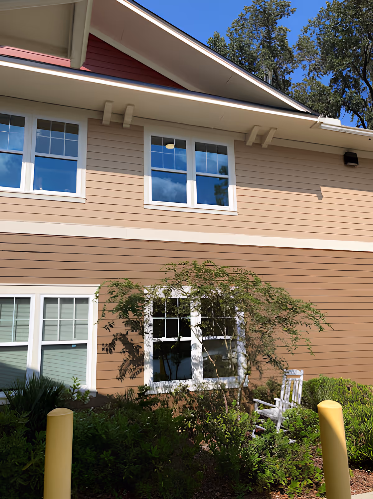 Exterior view of a beige and red two-story building with white-framed windows, surrounded by green shrubs and a small tree. A white rocking chair is placed near the window, and two yellow bollards are visible in the foreground under a clear blue sky.
