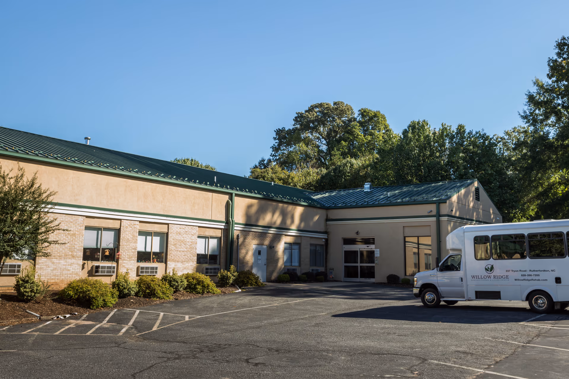 Exterior view of Willow Ridge Rehabilitation & Living Center building with a green metal roof, beige walls, several windows, and a parking lot in front. A white shuttle bus with the facility's name and contact information is parked on the right side. Trees and clear blue sky are visible in the background.