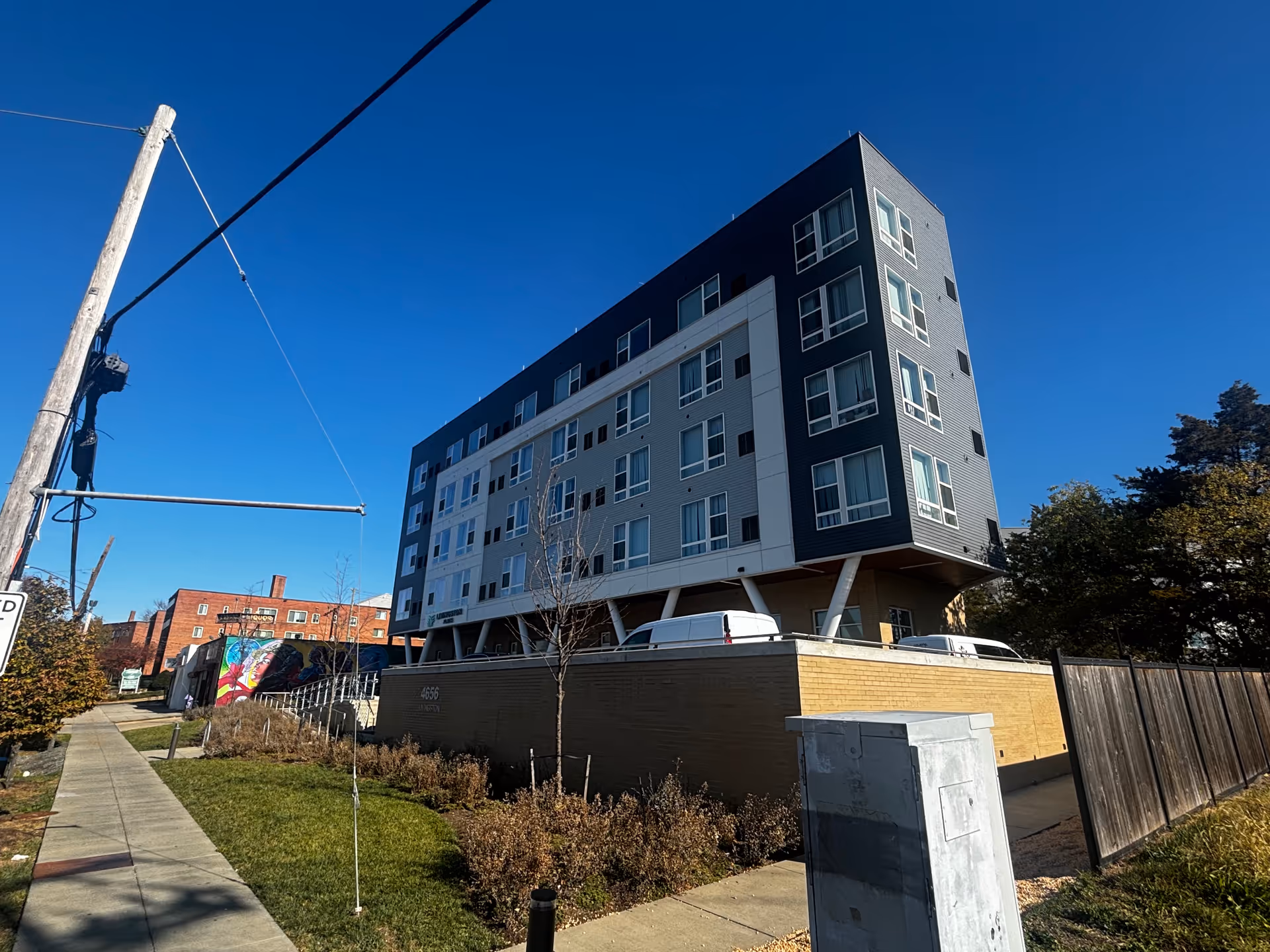 Exterior view of a modern multi-story building with large windows under a clear blue sky. The building is elevated on angled supports above a brick base, with a sidewalk, grass, and some trees in the foreground. A colorful mural is visible on a nearby wall to the left.