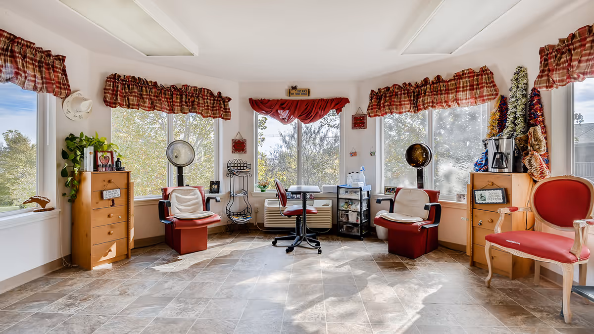 Bright interior salon/activity room with large windows, several red salon chairs and hooded hair dryers, small wooden cabinets and a central rolling chair.