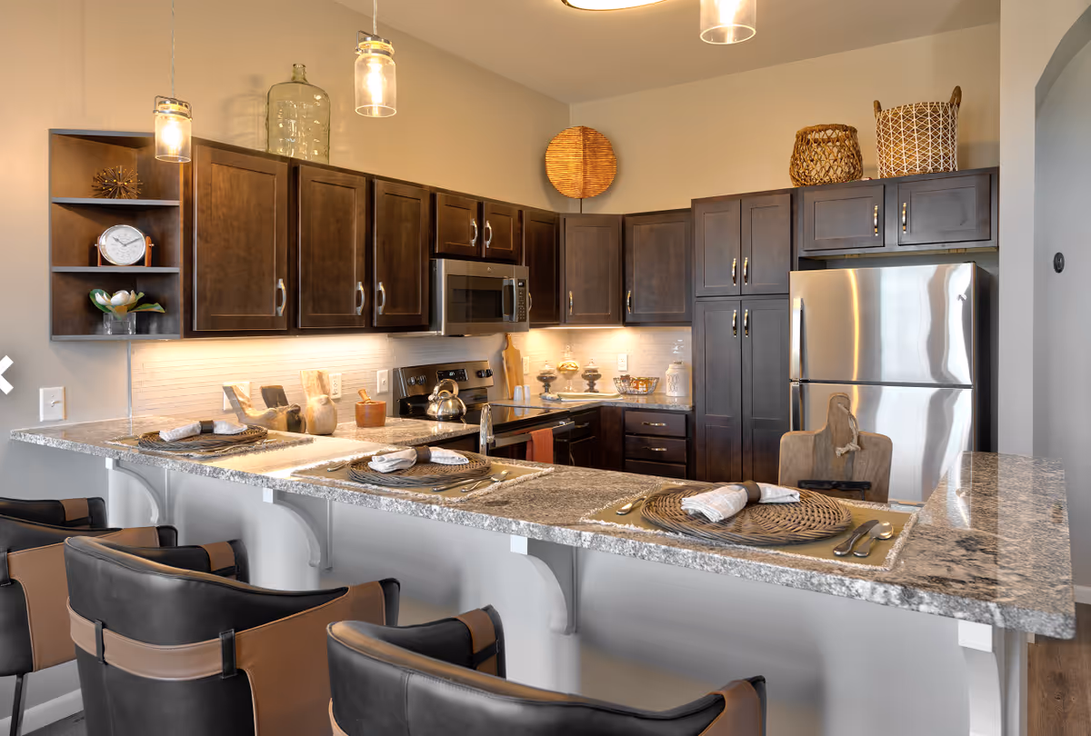 Modern kitchen with dark wooden cabinets, stainless steel appliances including a refrigerator and microwave, granite countertops with place settings, and three black and brown bar stools. Pendant lights hang above the counter, and decorative baskets and items are placed on top of the cabinets.