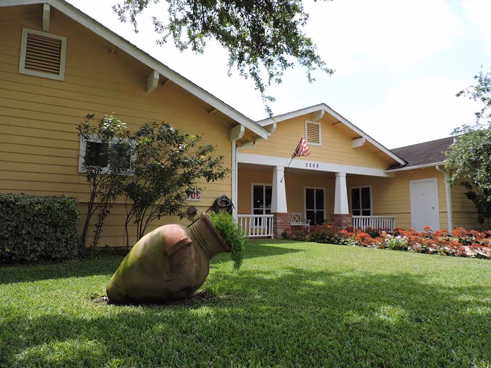 Front exterior of a yellow single-story assisted living building with a porch, American flag, lawn and a tilted large clay pot planter.