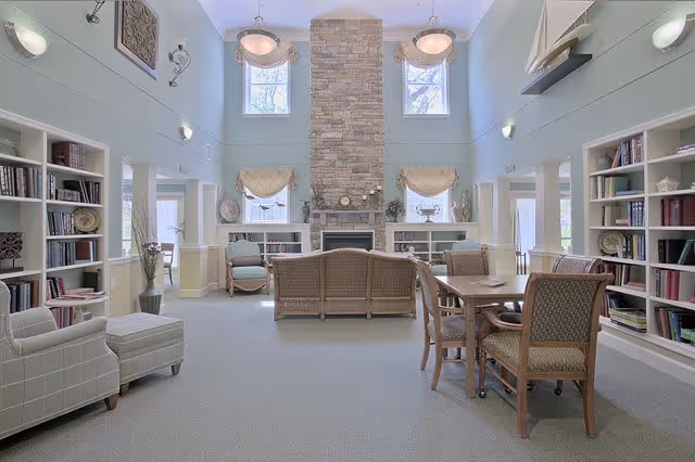 A spacious and well-lit common area with high ceilings and large windows. The room features a stone fireplace centered on the far wall, flanked by built-in bookshelves filled with books and decorative items. Comfortable seating includes a plaid armchair with an ottoman, a wicker sofa, and several wooden chairs around a table. The walls are painted light blue with white trim, and there are wall sconces and hanging light fixtures providing additional lighting.