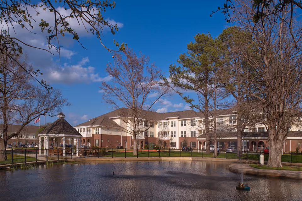 Exterior view of Crestview Retirement Community showing a large multi-story building with a brick and cream facade, surrounded by trees and a pond with a small fountain. There is a white gazebo near the pond and a clear blue sky with some clouds overhead.