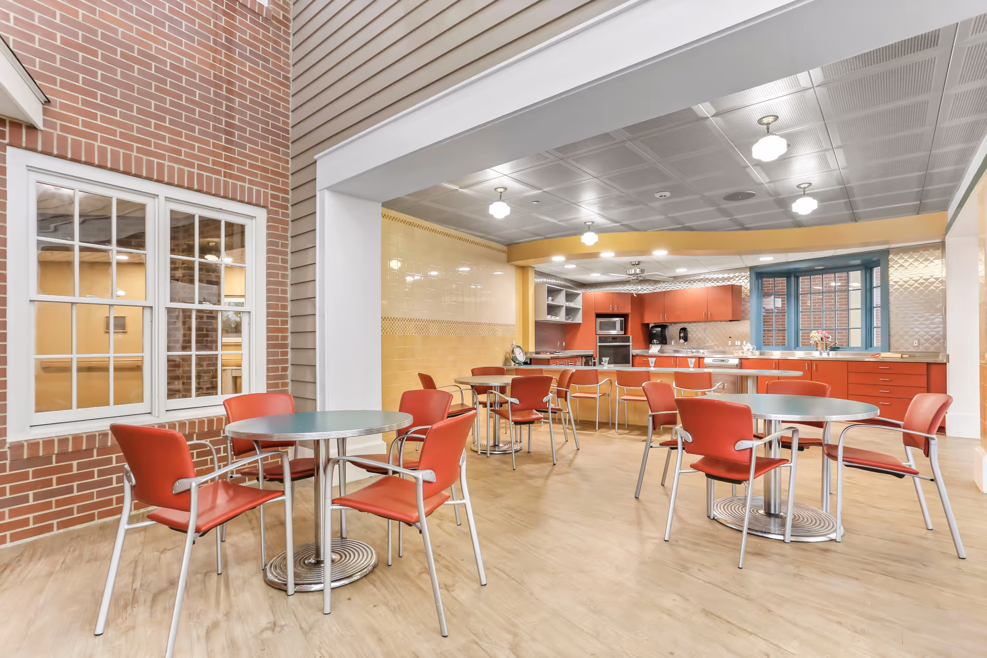 A bright and clean communal dining area with several round tables and red chairs. The space includes a kitchen area with red cabinets, a microwave, and a coffee maker. The room has a mix of brick and siding walls, with large windows and ceiling lights providing ample illumination.