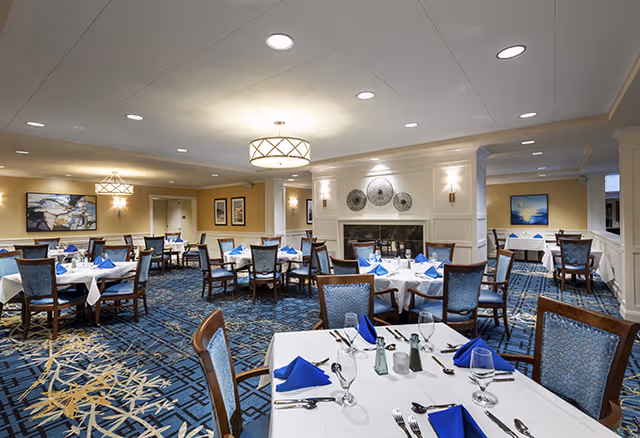 A spacious dining room with multiple tables covered in white tablecloths and set with blue napkins, silverware, glasses, and salt and pepper shakers. The room features blue patterned carpet, elegant wooden chairs with blue upholstery, a fireplace with decorative plates above it, and soft overhead lighting. Artwork is displayed on the walls.