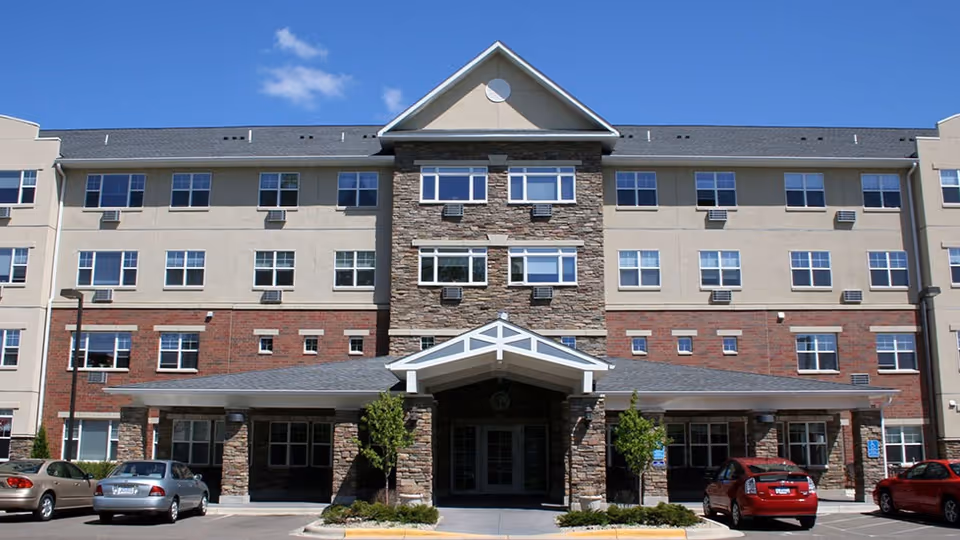 Front exterior view of a multi-story senior living facility building with a covered entrance, stone and brick facade, multiple windows, and several parked cars in front under a clear blue sky.