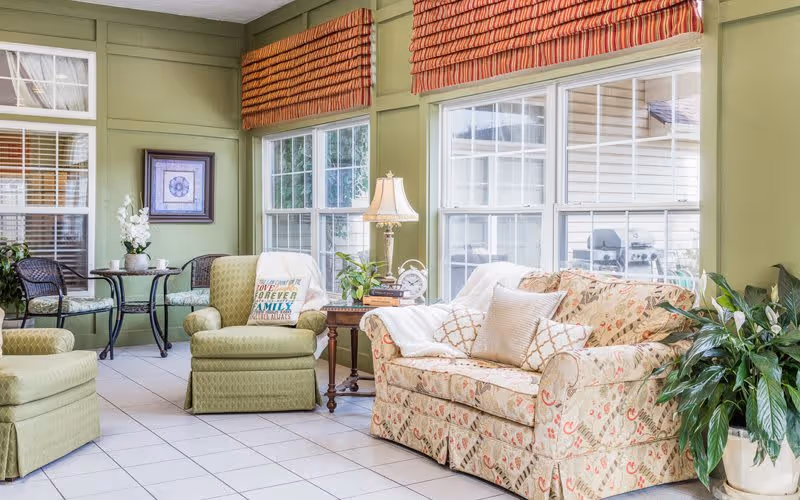 Sunlit common area with a floral sofa, green upholstered chairs, side tables and large windows with striped valances.