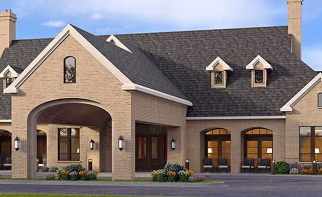 Front exterior view of a senior living facility building with beige brick walls, multiple windows, a covered entrance with an archway, and a dark shingled roof under a blue sky.
