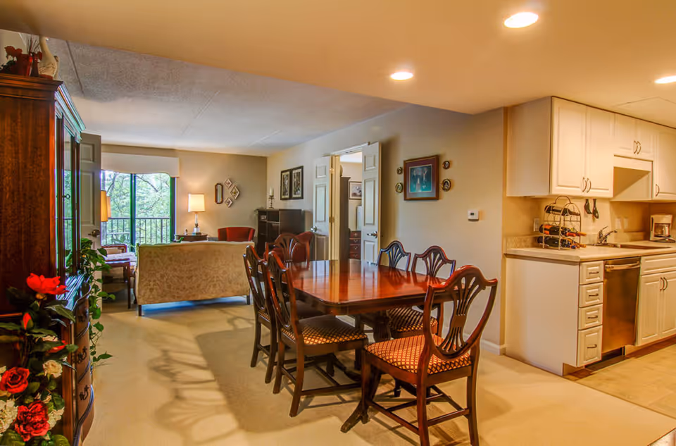 Interior view of a senior living facility showing a dining area with a wooden table and six chairs, adjacent to a kitchen with white cabinets and stainless steel dishwasher. In the background, there is a living room area with a sofa, armchair, lamp, and large window with a view of greenery outside. The room is warmly lit with ceiling lights.
