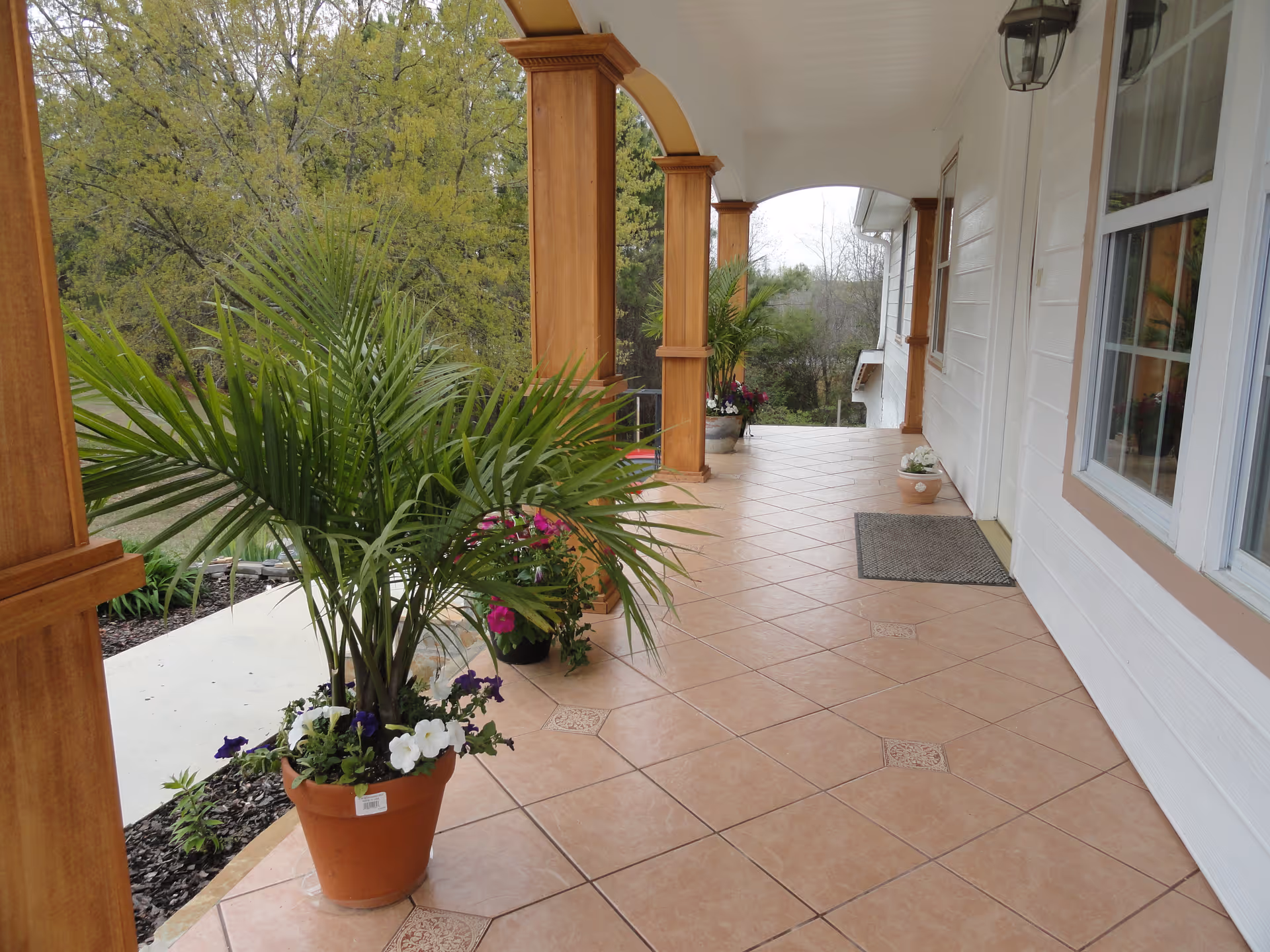 A covered outdoor porch area with tiled flooring and wooden columns. Several potted plants with green leaves and colorful flowers are placed along the edge of the porch. The porch overlooks a garden area with trees and greenery in the background.