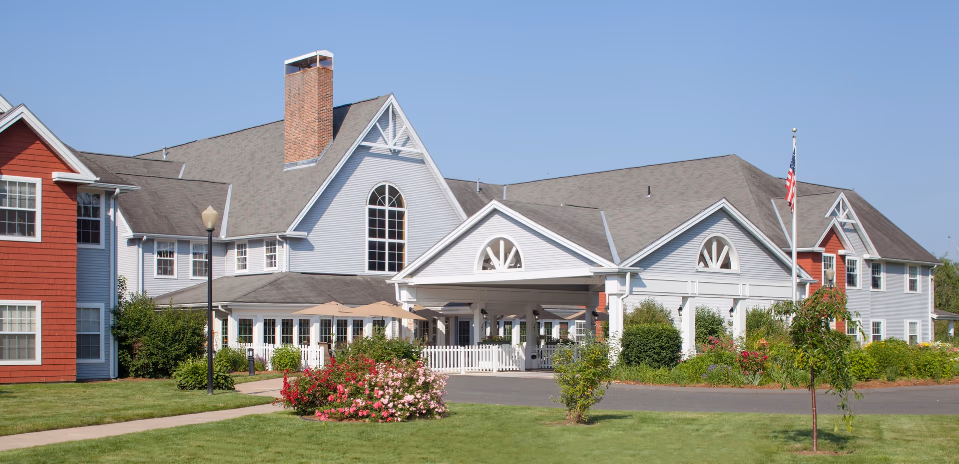 Exterior view of a large senior living facility building with gray and red siding, multiple peaked roofs, a brick chimney, and a covered entrance. The foreground features a well-maintained lawn, flowering bushes, a sidewalk, and an American flag on a flagpole.