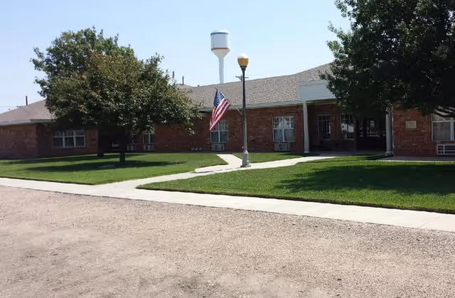Single-story brick building with a grassy front lawn, an American flag on a lamppost, and a water tower visible behind.