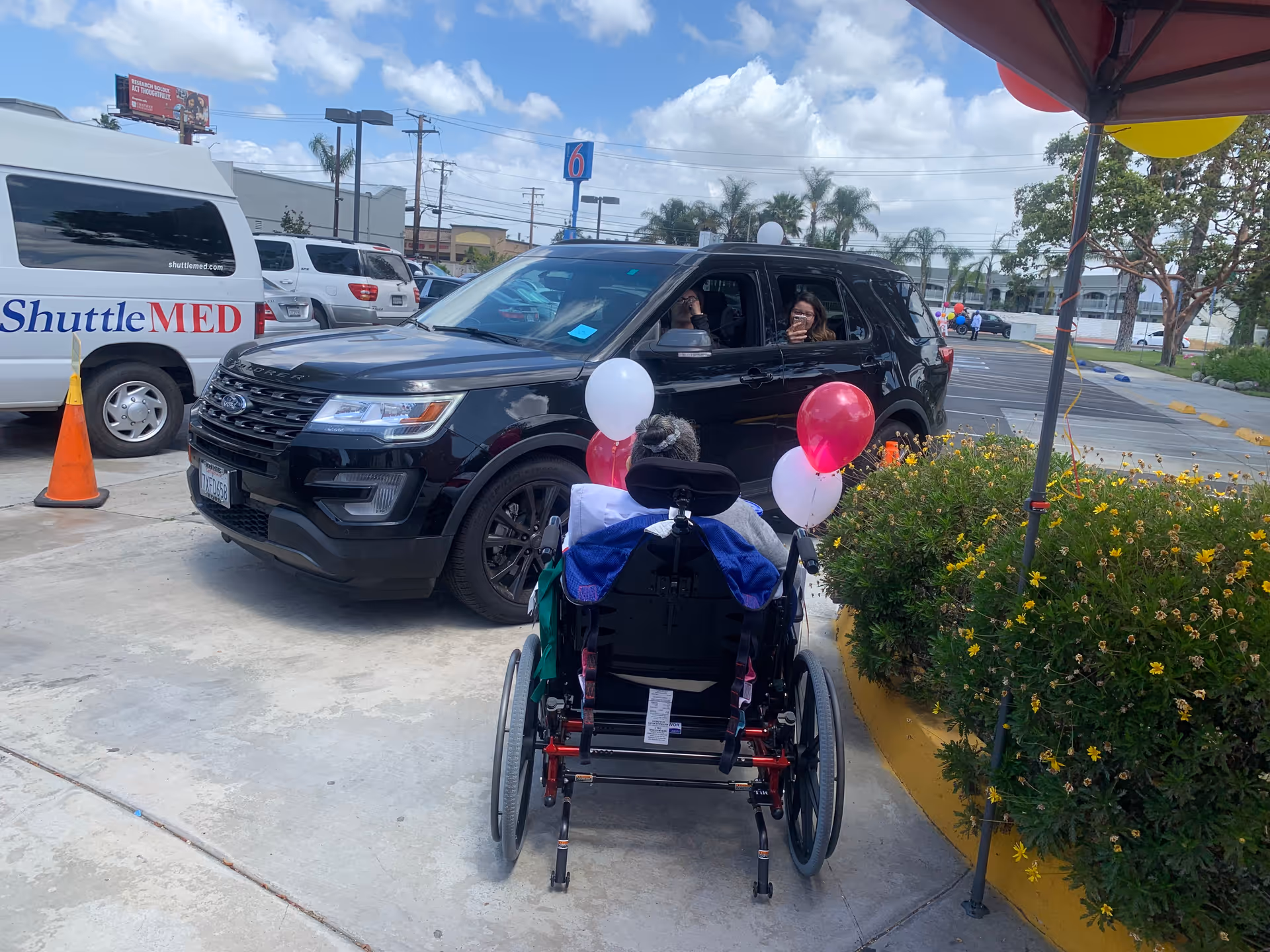 A person in a wheelchair decorated with red and white balloons is outside near a black SUV with two people inside. The scene is in a parking lot with other vehicles, including a white van labeled ShuttleMED. There are yellow flowers and a canopy with balloons on the right side.