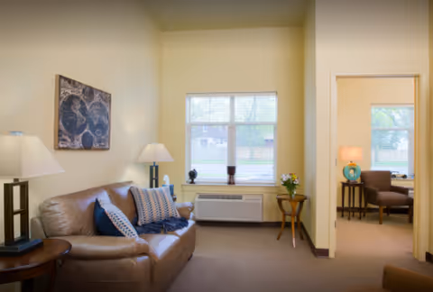 A cozy living room area with a brown leather sofa adorned with patterned pillows and a throw blanket. There are two table lamps on either side of the sofa, a window with blinds letting in natural light, and a small round table with a flower vase. An adjoining room with a chair and a lamp is visible through an open doorway.