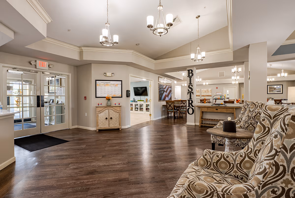 Interior view of a senior living facility lobby area with patterned armchairs and a small round table in the foreground. The space features wood flooring, beige walls, and multiple ceiling light fixtures. A column with the word 'BISTRO' in large letters is visible near a dining area with tables and chairs. Double glass doors with an exit sign are on the left side, and a bulletin board labeled 'Life Enrichment' is mounted on the wall.