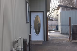 Side exterior of a building showing a white oval-glass door, a small window, a chain-link gate, and a nearby shed.