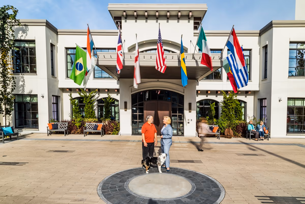 Two elderly people standing and talking in front of the entrance to a building with multiple international flags hanging above. There is a dog with them. The building has large windows and benches with cushions on either side of the entrance. Other people are sitting on benches in the background.
