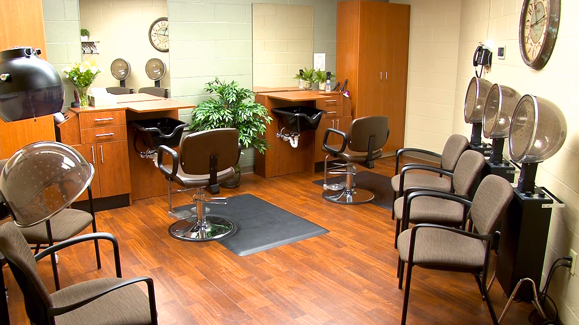 Interior of a senior living facility hair salon with wooden flooring, two styling chairs in front of sinks, three hair drying chairs with hooded dryers, a clock on the wall, and several waiting chairs.