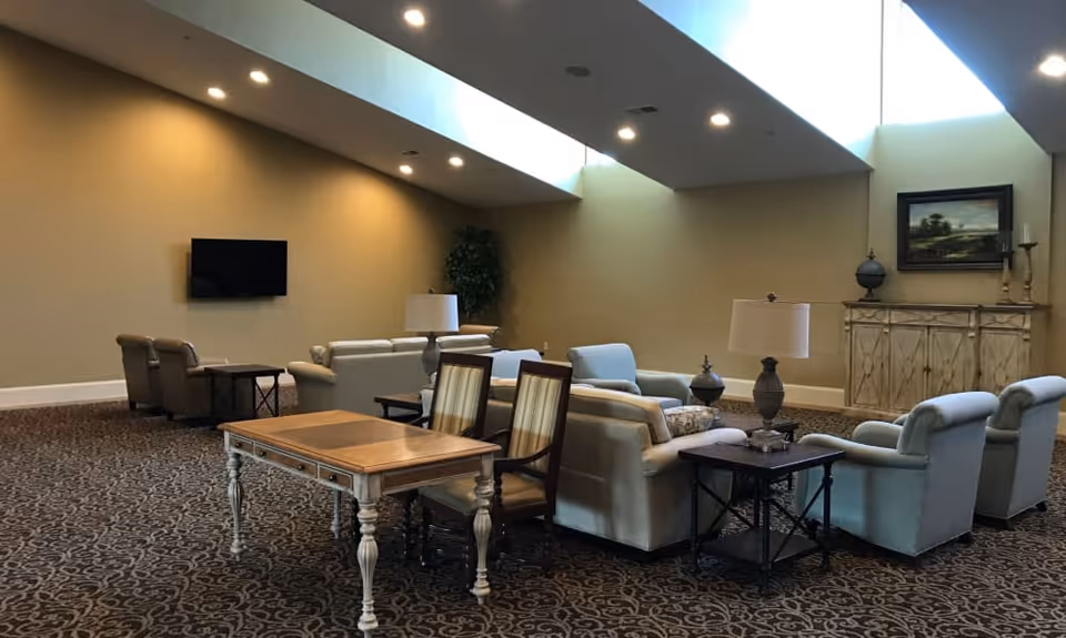 A spacious living room area with multiple seating options including sofas and armchairs arranged around side tables with lamps. There is a wooden table with two chairs in the foreground. The room has high ceilings with recessed lighting and skylights, beige walls, a mounted flat-screen TV, a large plant in the corner, and a decorative cabinet with a painting and candle holders on top.