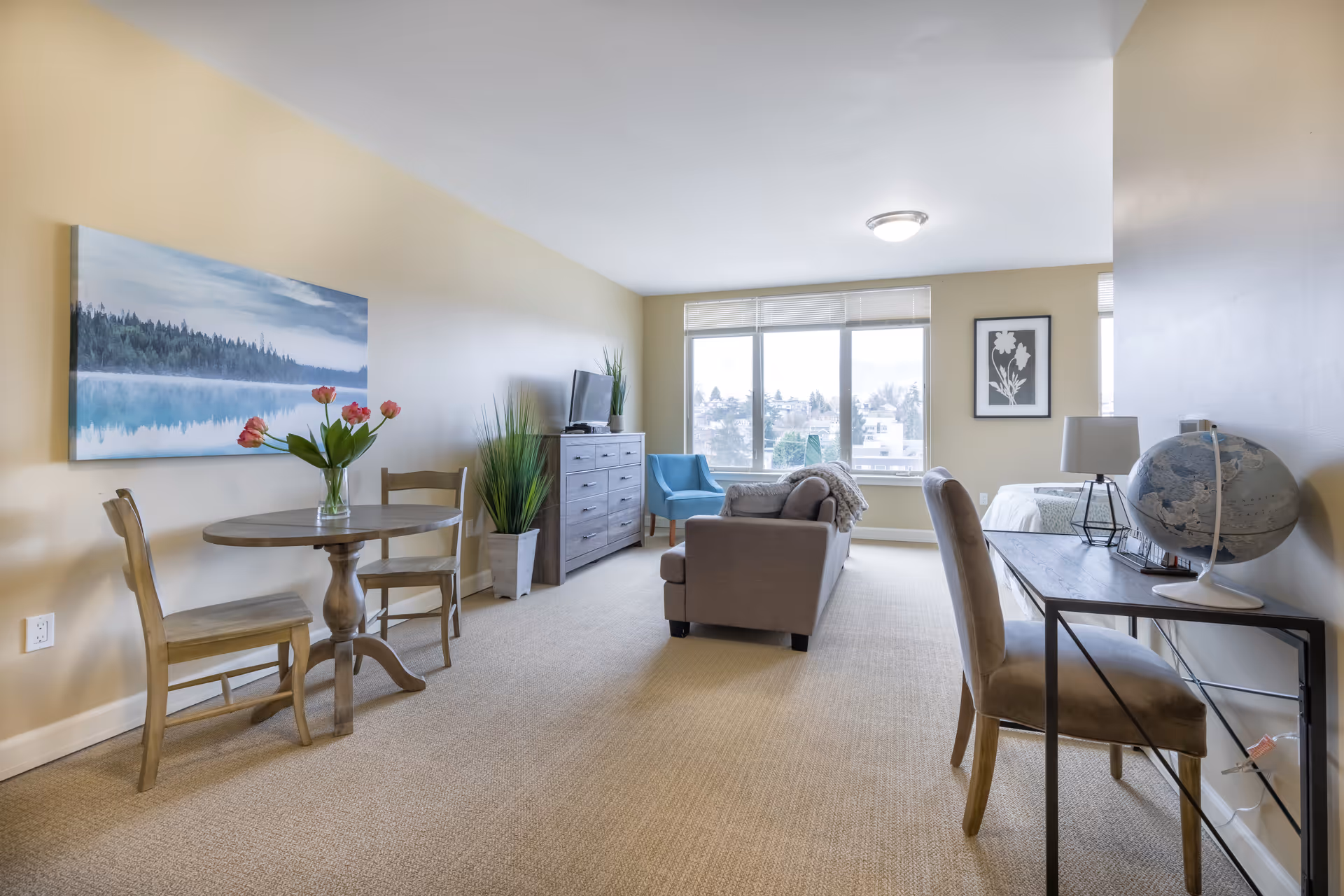 A bright and spacious living area in a senior living facility with beige walls and carpet. The room features a small round wooden dining table with two chairs and a vase of pink flowers, a gray sofa, a blue armchair, a wooden dresser with a TV on top, and a desk with a chair and a globe. Large windows let in natural light and offer a view of the outside.
