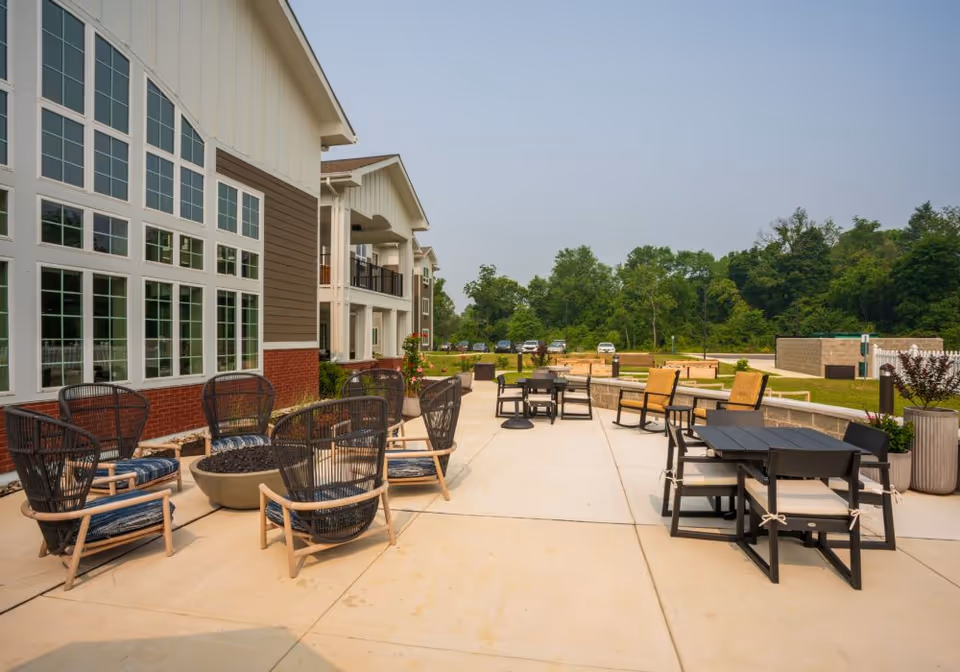 Outdoor patio area at Juniper Village at Newark with multiple seating arrangements including black wicker chairs with cushions around a fire pit, and black tables with chairs. The patio is adjacent to a building with large windows and surrounded by greenery and trees in the background.