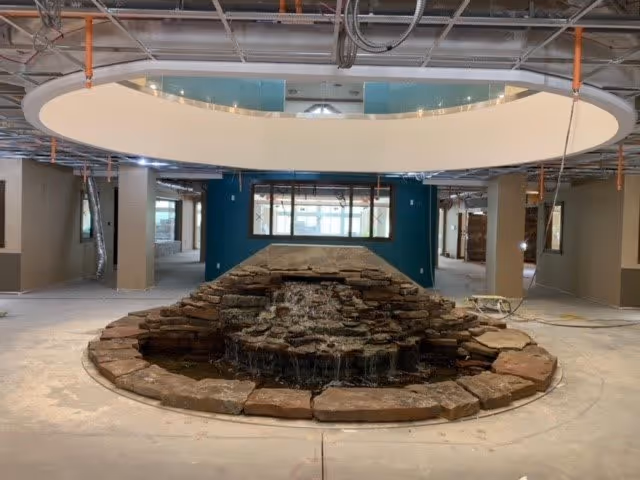 Indoor area under construction featuring a stone water fountain in the center with water cascading down multiple tiers. Above the fountain is a large circular ceiling feature with recessed lighting. The space has exposed ceilings with visible ductwork and wiring, and walls painted in neutral tones with a teal accent wall in the background.