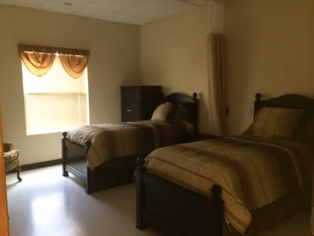 A bedroom with two single beds featuring dark wooden frames and matching striped bedding. There is a window with a brown valance letting in natural light, a small dark wooden cabinet between the beds, and a chair partially visible on the left side.