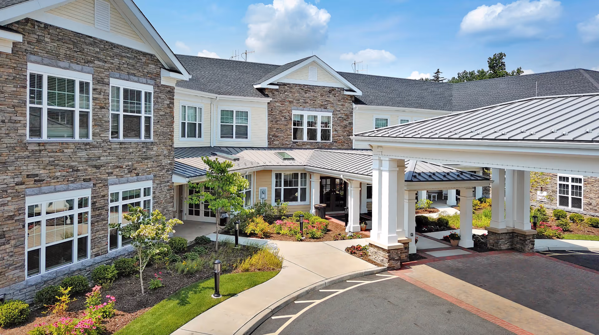 Front entrance of a two-story stone-clad senior living building with a covered porte-cochere and landscaped driveway.