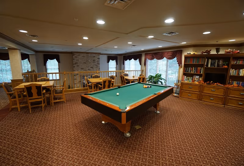 A spacious common area with a pool table in the center, surrounded by wooden tables and chairs. The room has carpeted floors with a patterned design, large windows with maroon valances, and a wooden bookshelf filled with books and decorative items. The ceiling has recessed lighting and there is a railing separating the area from a lower level.