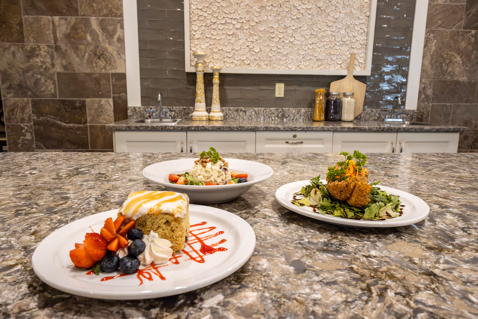 Three plates of food arranged on a granite countertop in a kitchen setting. The dishes include a dessert with strawberries and blueberries, a salad with a creamy topping and nuts, and a salad with greens and a fried item garnished with parsley. The background shows a kitchen counter with two sinks, decorative jars, and a wall adorned with a textured art piece.