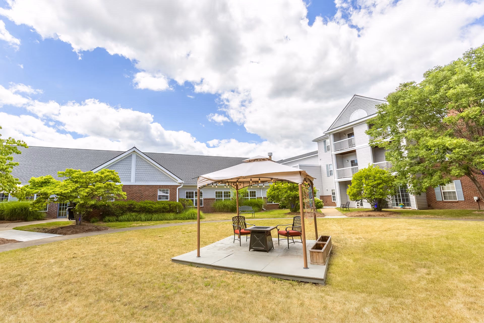 Outdoor courtyard area at Independence Village of White Lake featuring a small gazebo with a canopy, four chairs, and a central fire pit table on a concrete slab surrounded by grass and trees, with residential buildings in the background under a partly cloudy sky.