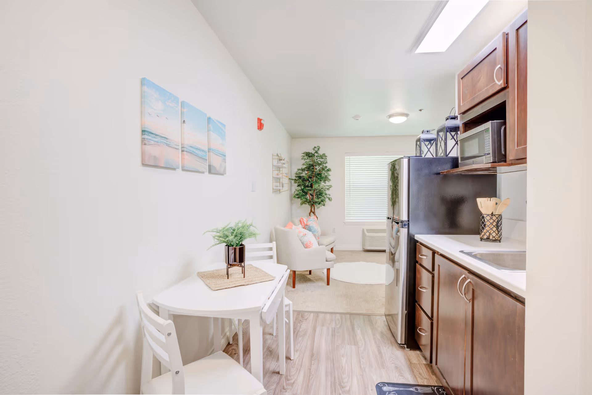 A bright and clean interior space featuring a small kitchen area with dark wood cabinets, a stainless steel refrigerator, and a microwave. Adjacent to the kitchen is a small white dining table with two chairs and a potted plant on top. In the background, there is a cozy sitting area with a white armchair, colorful pillows, a round white rug, a tall green plant, and a window with blinds letting in natural light. The walls are white with a three-panel beach-themed artwork hanging above the dining table.