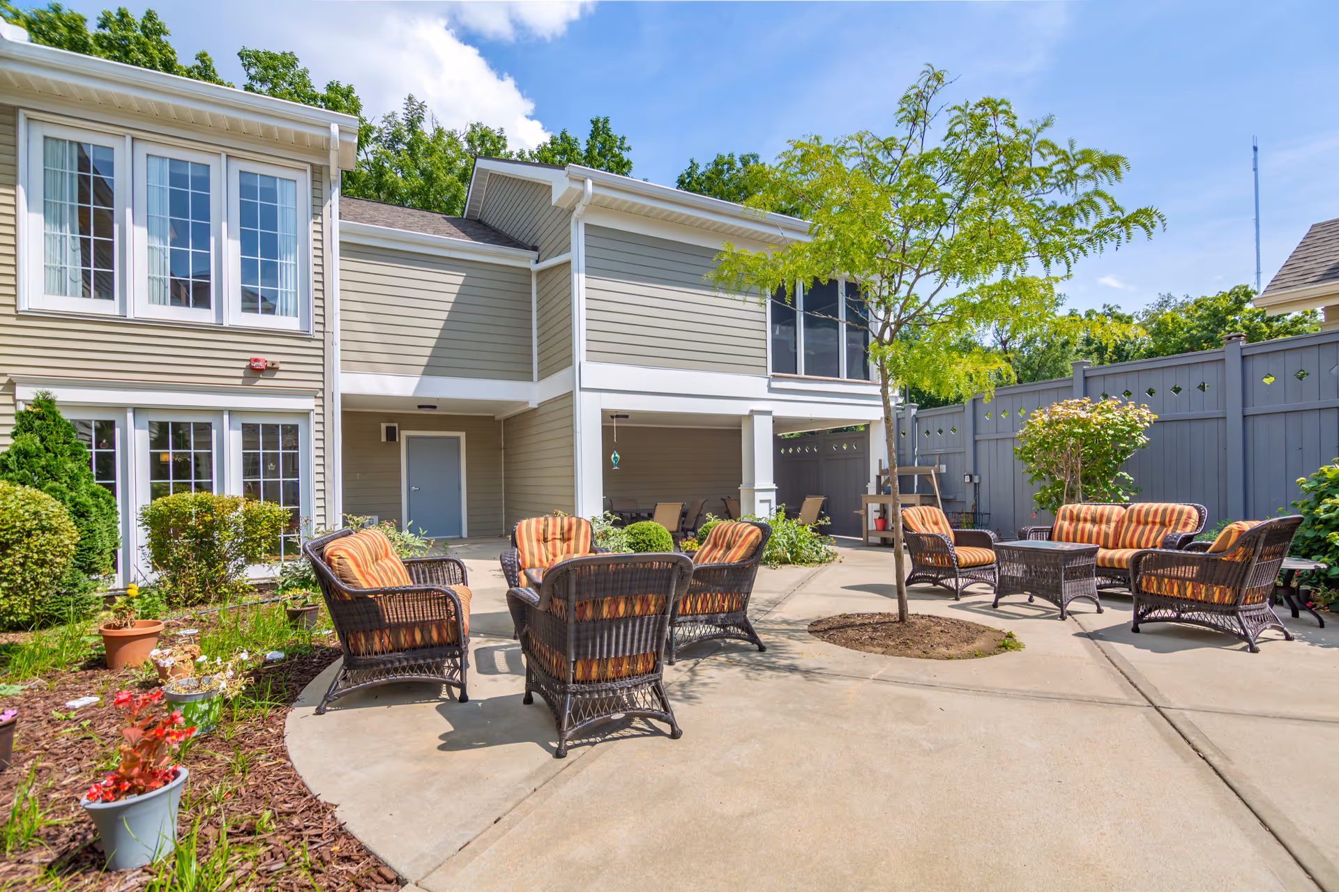 Outdoor patio area at Brookdale Madison West with wicker chairs and sofas featuring orange striped cushions arranged around small tables. The patio is surrounded by a gray fence and landscaped with small trees, bushes, and potted plants. The building exterior is beige with white trim and large windows under a blue sky with some clouds.