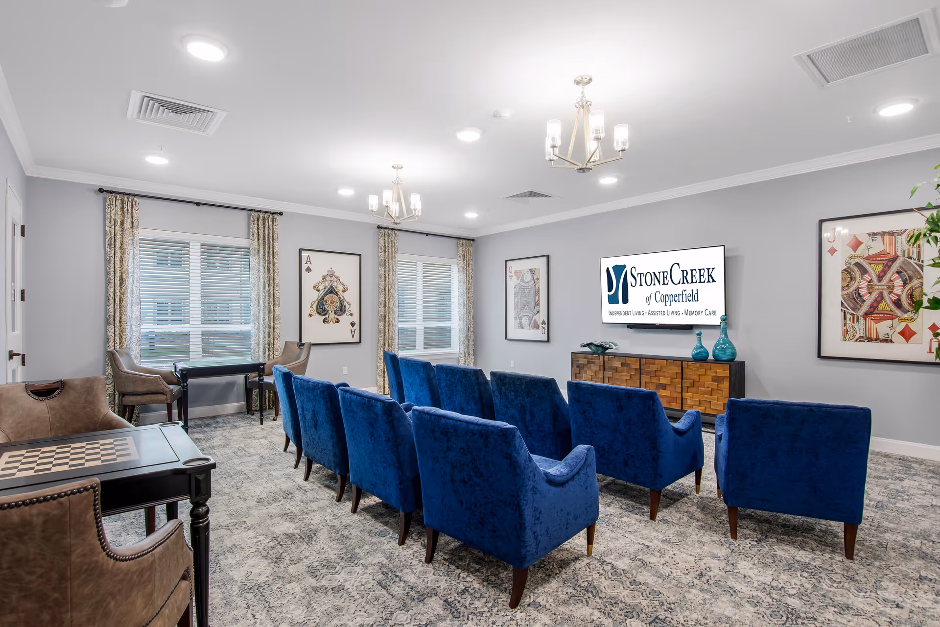 A bright senior living common room with rows of blue upholstered chairs facing a wall-mounted TV showing the StoneCreek of Copperfield logo.
