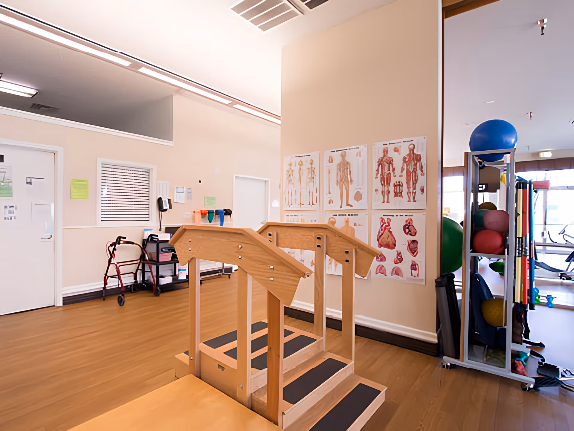 Physical therapy room with a wooden rehabilitation staircase in the center, anatomical charts on the wall, a walker near the door, and exercise equipment including stability balls and resistance bands on a rack.