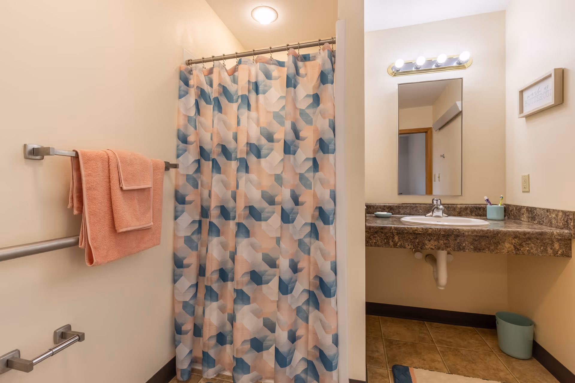 Bathroom with a shower area covered by a geometric patterned shower curtain in shades of blue, peach, and white. On the left wall, there are two peach-colored towels hanging on a metal towel rack. To the right, there is a countertop with a sink, a mirror above it with four lights, a toothbrush holder with toothbrushes, and a small soap dish. The floor is tiled, and there is a small trash bin under the counter. A small framed sign is mounted on the wall above the countertop.