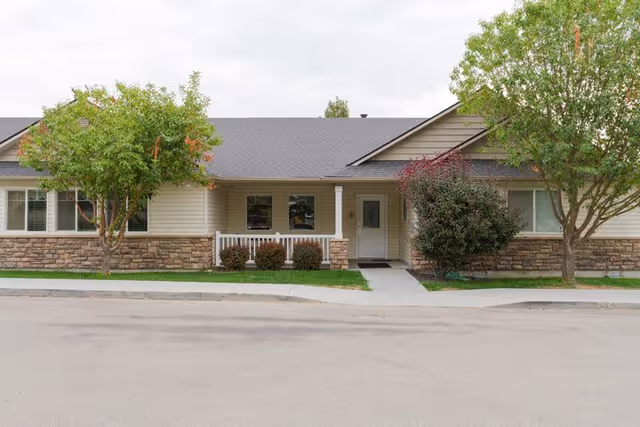 Single-story residential building with beige siding and stone accents, a gray shingled roof, a small front porch with white railing, and two trees on either side of the entrance.