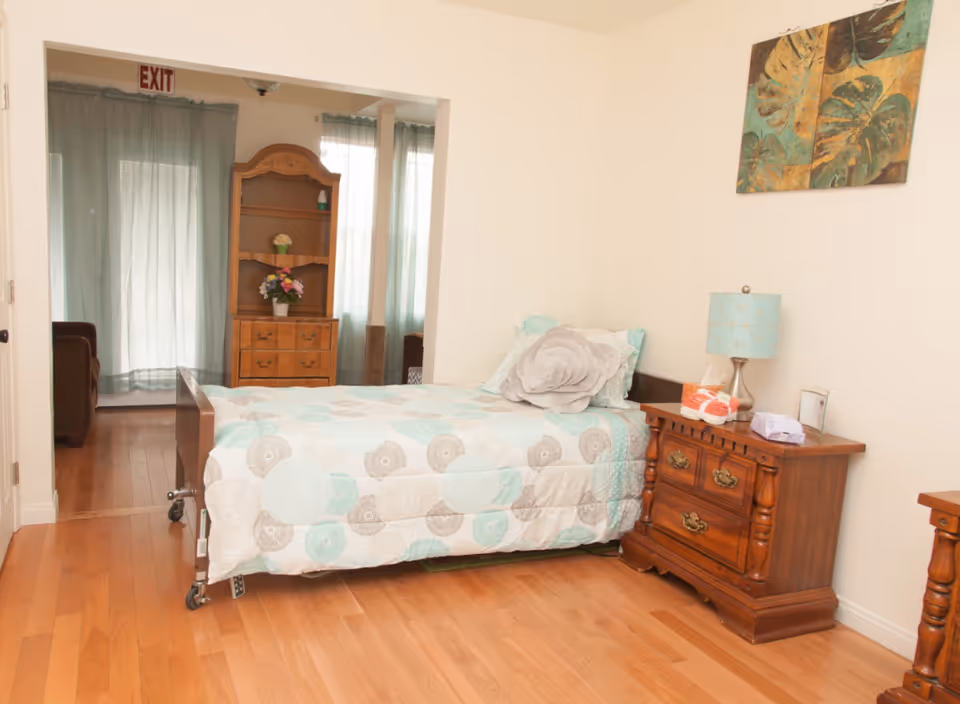 A bedroom with a single bed covered in a light blue and beige patterned quilt, a large flower-shaped pillow, and a wooden nightstand with a blue lamp, tissue box, and other small items. There is a wooden dresser with shelves and flowers in the background, light blue curtains, and an exit sign above the doorway.